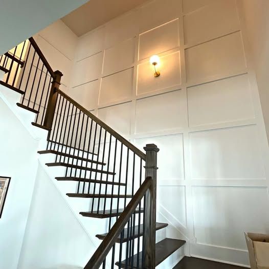 Staircase with dark railing in a bright foyer, beside white paneled walls and a lit wall sconce