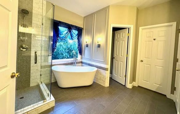 A modern bathroom features a glass shower, a freestanding white soaking tub by a window, and two white doors.