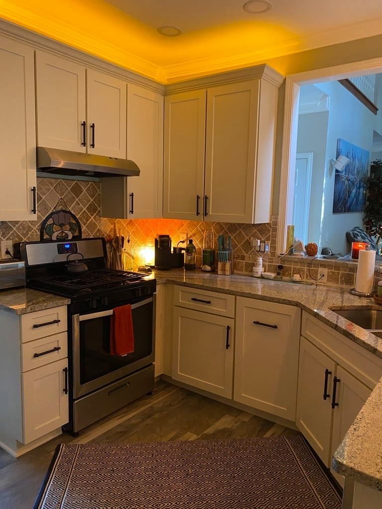 Small kitchen with white cabinets, black stove, warm lighting, and a granite countertop with items on it
