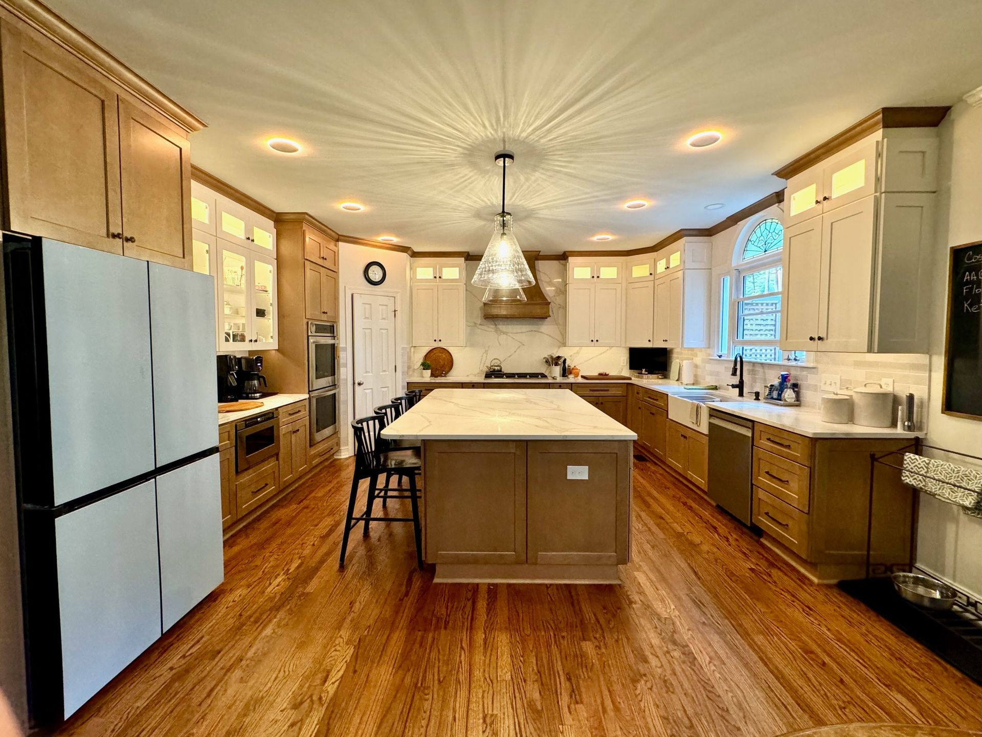 Bright kitchen with wood floors, white cabinets, stainless fridge, and a large island under pendant lights.