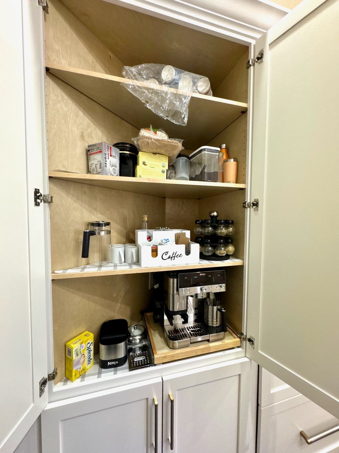 Open pantry cabinet with shelves holding kitchen appliances, jars, mugs, and storage items.