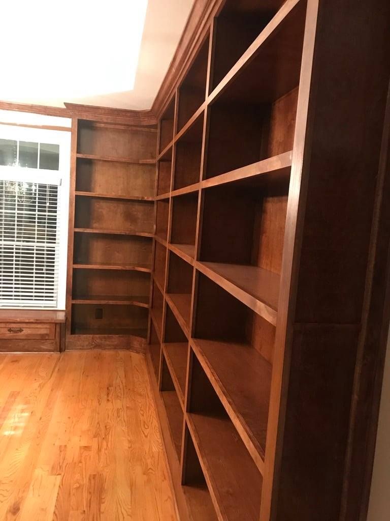 Empty room with built-in wooden bookshelves and hardwood floors, lit by a window on the left.