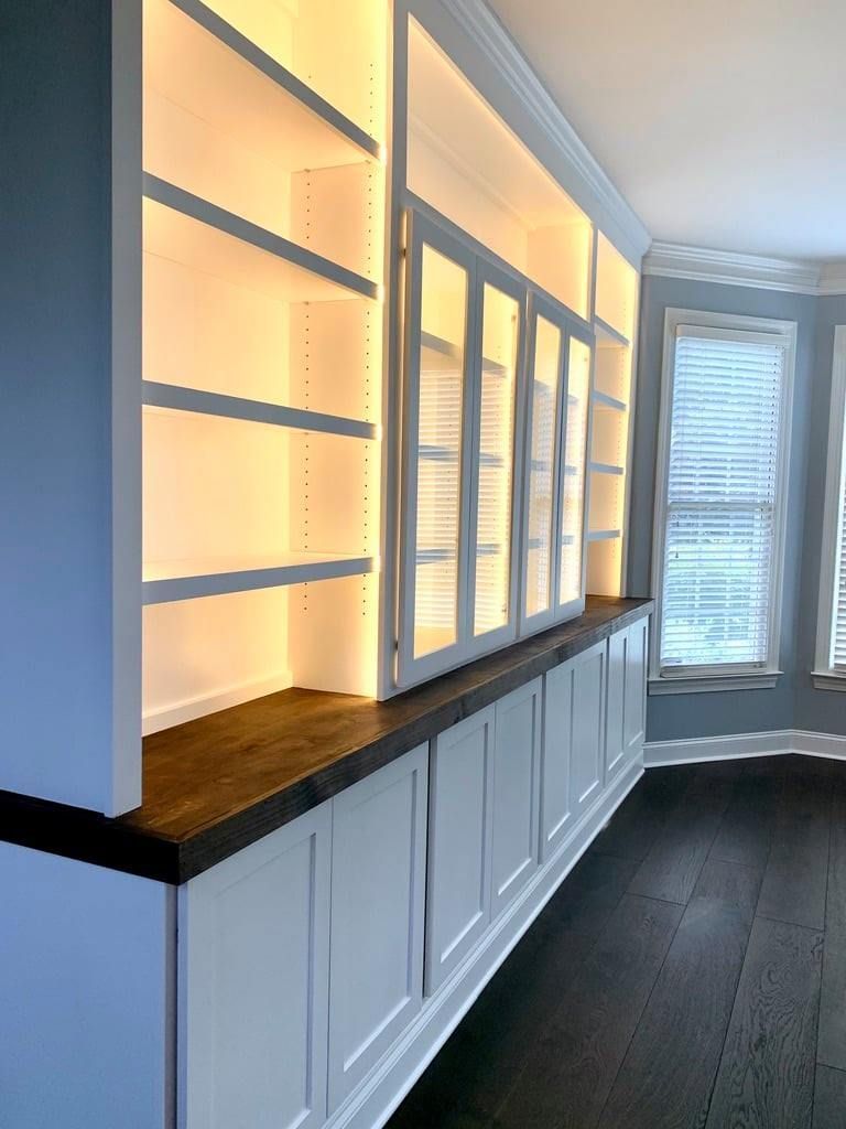 Built-in lit shelving and cabinets along a dark hallway with wood countertops and windows.