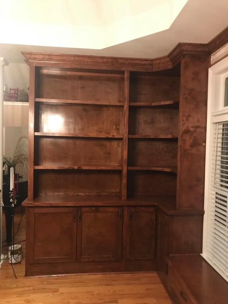 Built-in wooden bookshelf with cabinets in a living room corner, empty shelves beside a window with blinds