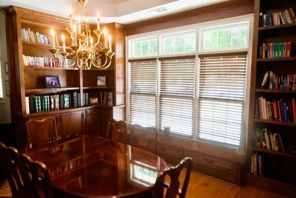 Warm wood-paneled home office with bookshelves, chandelier, and large window with blinds