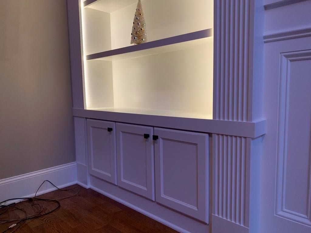 Built-in white shelving unit with under-shelf lighting above cabinet doors in a room corner.