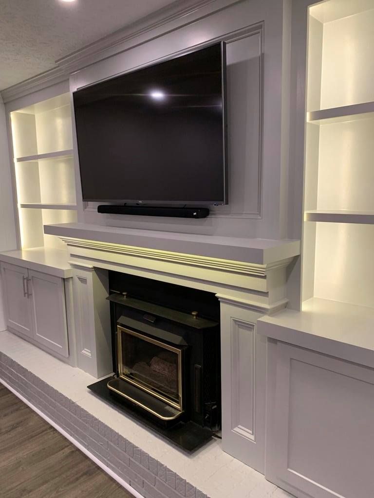 Built-in wall unit with TV above a fireplace, gray cabinetry, and lit display shelves