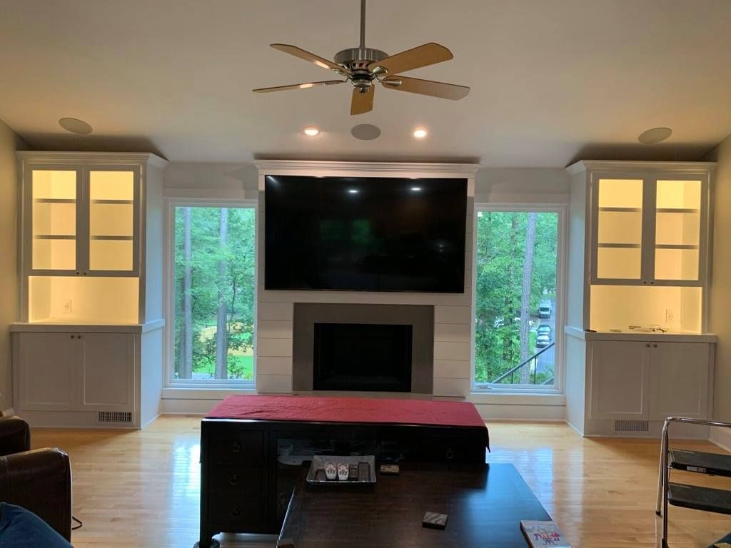 Living room with a large TV, fireplace, built-in shelves, windows, and a red ottoman centerpiece