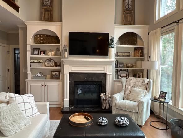 Bright living room with fireplace, TV, cream chairs, built-in shelves, and large windows