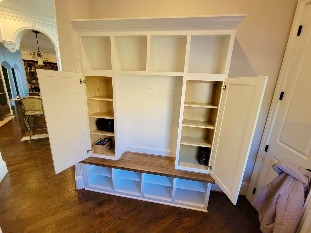 White built-in storage cabinet with open shelves and a wood bench, doors open in a hallway.