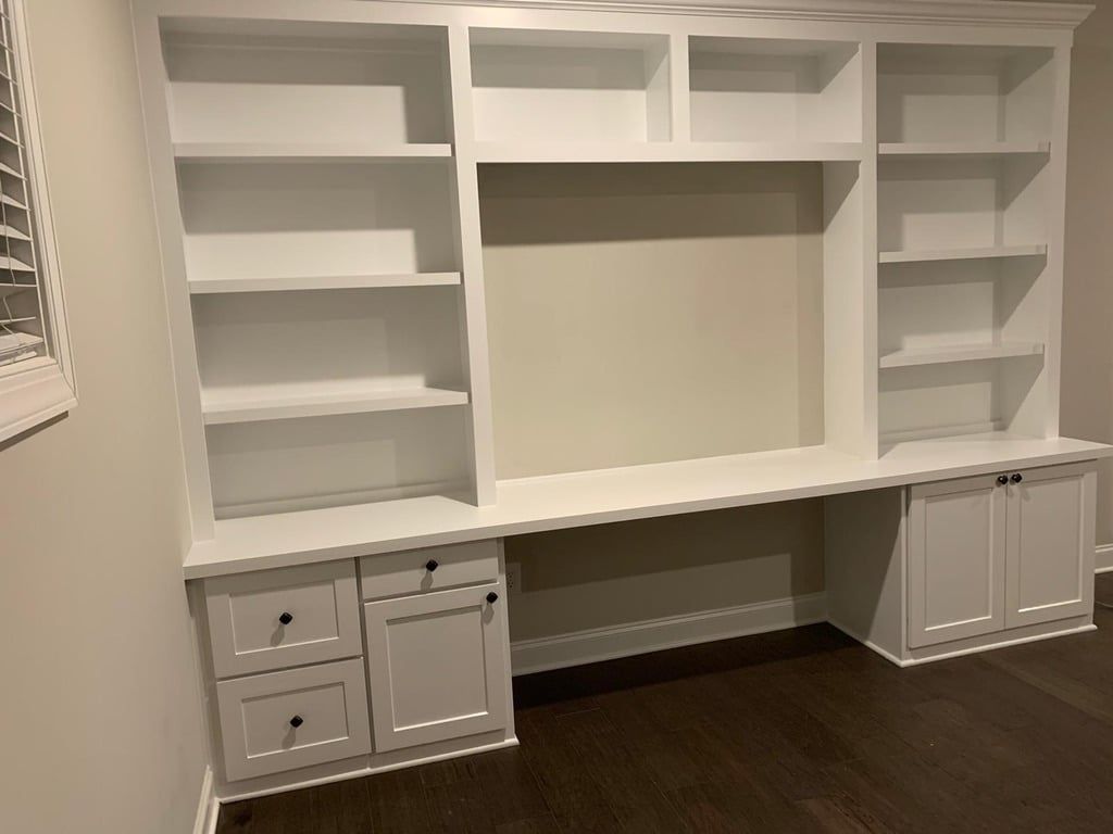 White built-in office shelving and cabinets with a central desk opening against a dark wood floor.