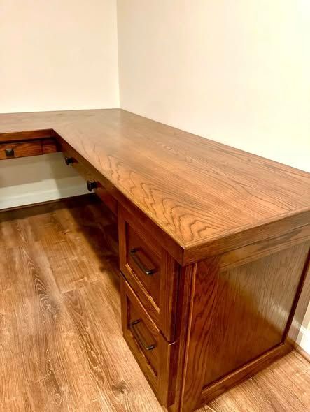 Wooden L-shaped desk with drawers in a corner against beige walls and wood flooring