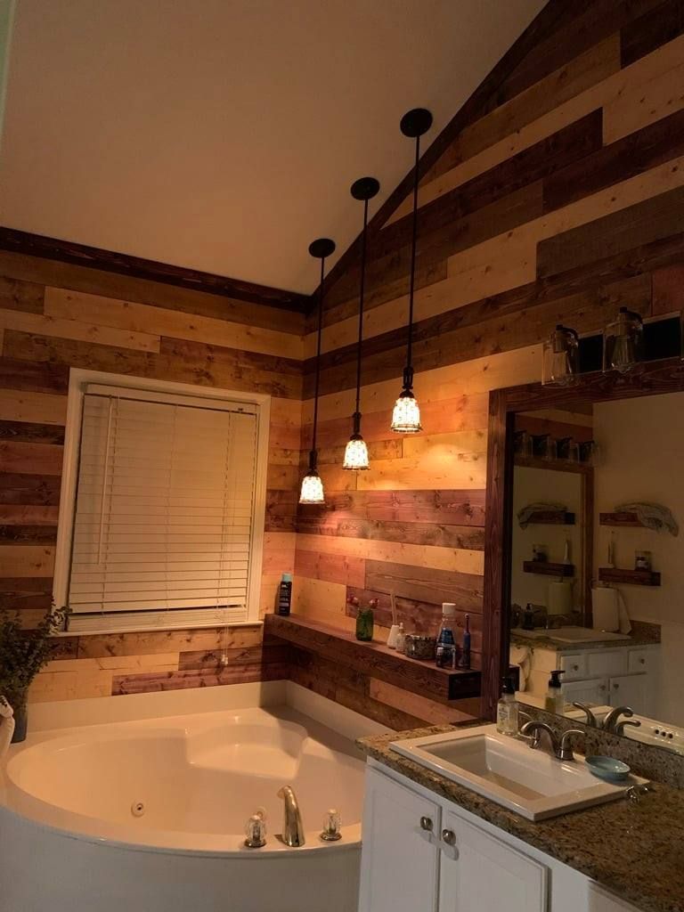 Warm bathroom with wood plank walls, vanity mirror, pendant lights, and a corner bathtub near a window blind