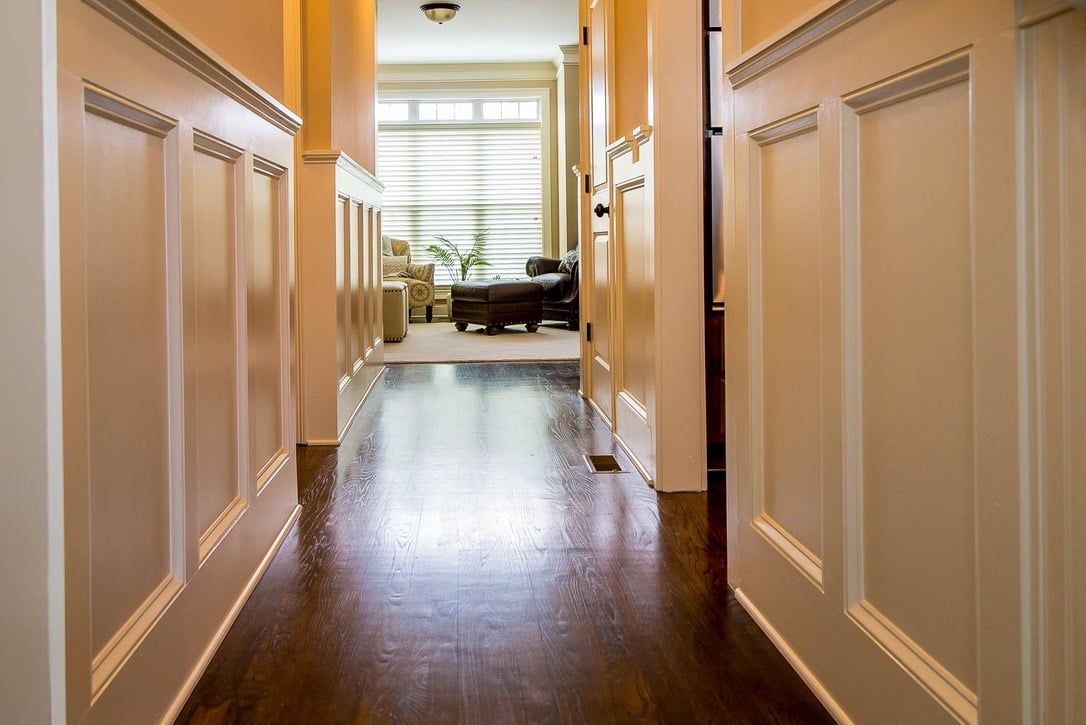 Narrow hallway with white walls and dark wood floor, leading to a bright sitting area with a sofa and window.