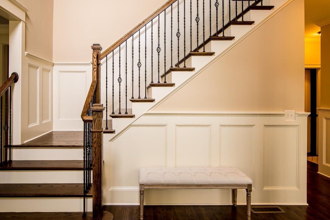 Interior staircase with dark railing, white paneled walls, and a wooden bench in a warmly lit hallway