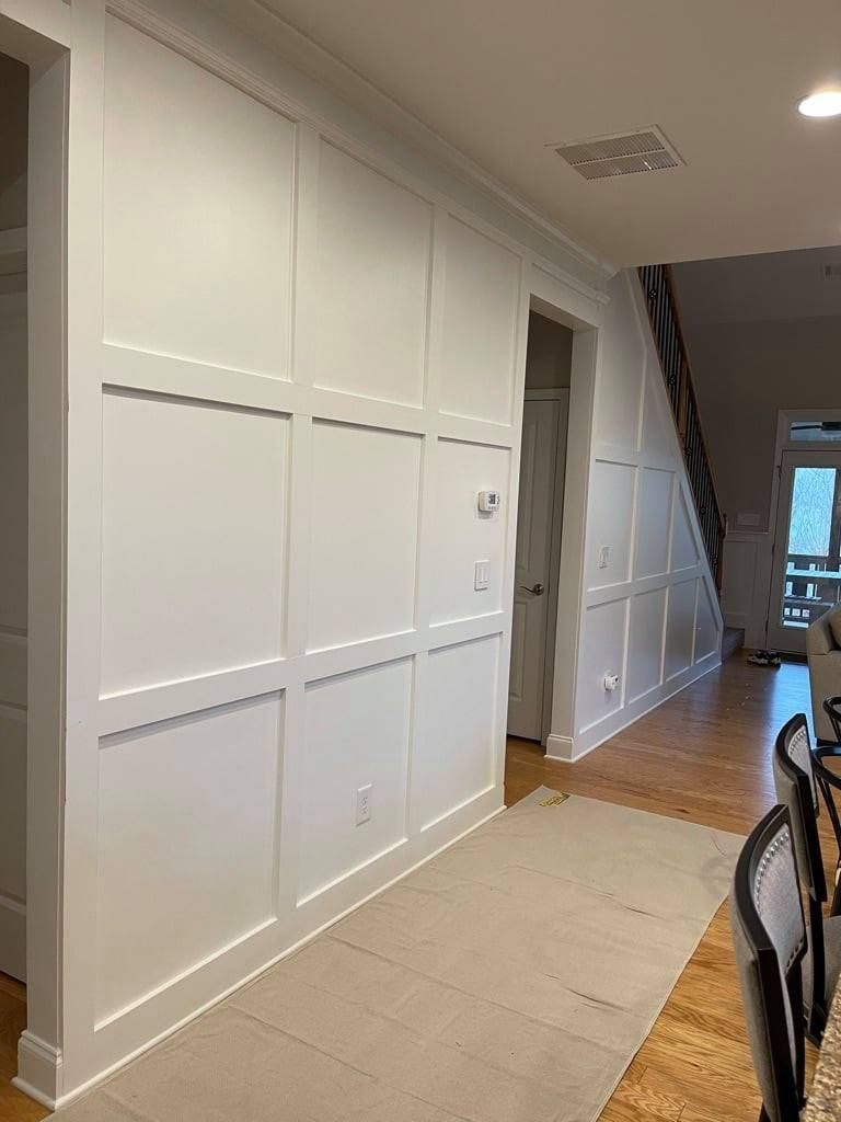 Bright hallway with white paneled wall, beige floor, and chairs along the right side.