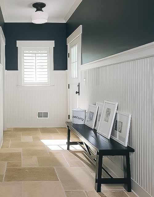 Bright hallway with white wainscoting, dark walls, a bench, framed art, and sunlight from a window.