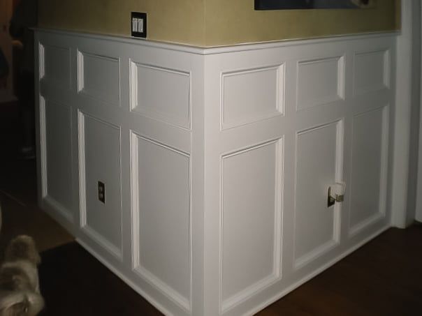 White wainscoted half wall in a corner of a room, with beige walls and dark flooring.