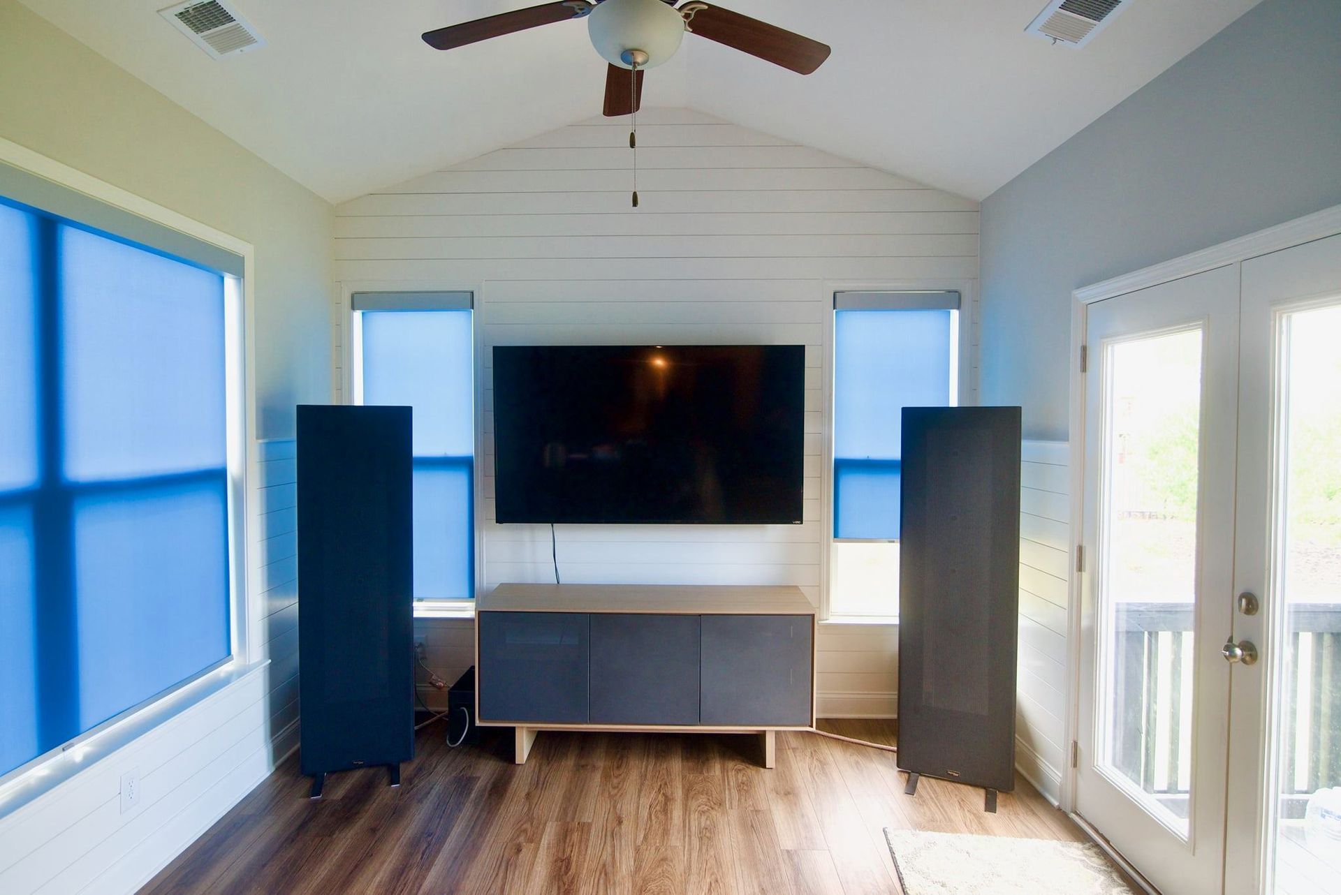 Bright living room with a wall-mounted TV, tall speakers, blue shades, and a ceiling fan