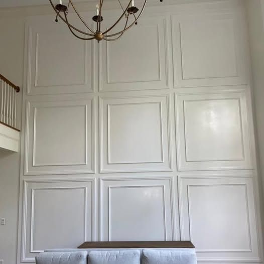 White paneled wall with three columns of recessed squares, a chandelier above, and a bench at the bottom.