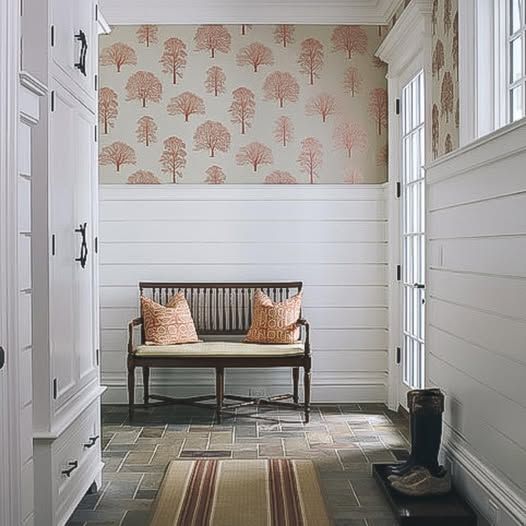 Narrow hallway with floral wallpaper, white paneling, a wooden bench with pillows, and rubber boots by the door.