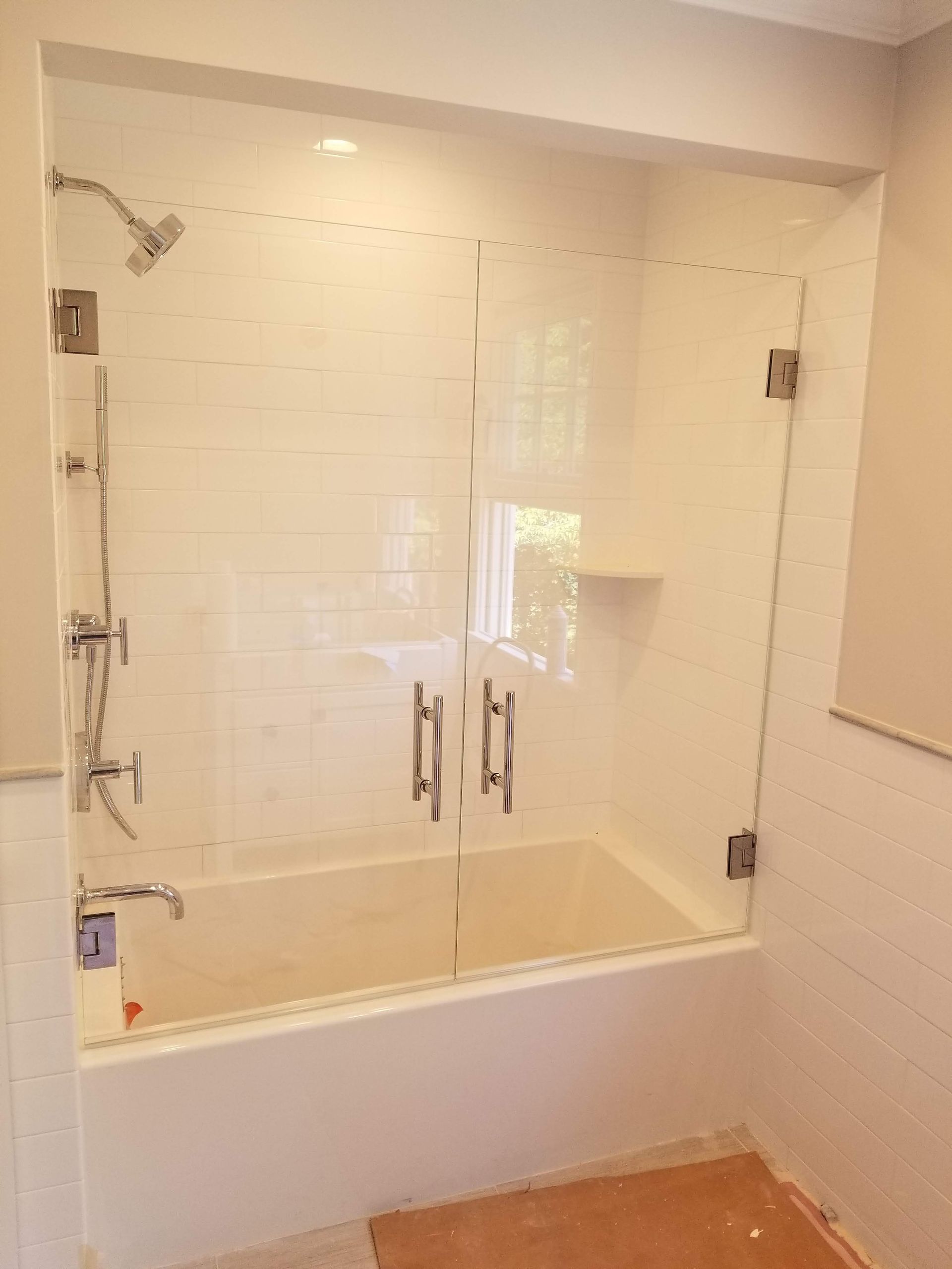 Glass-enclosed shower over a white bathtub with white tile walls and chrome fixtures.