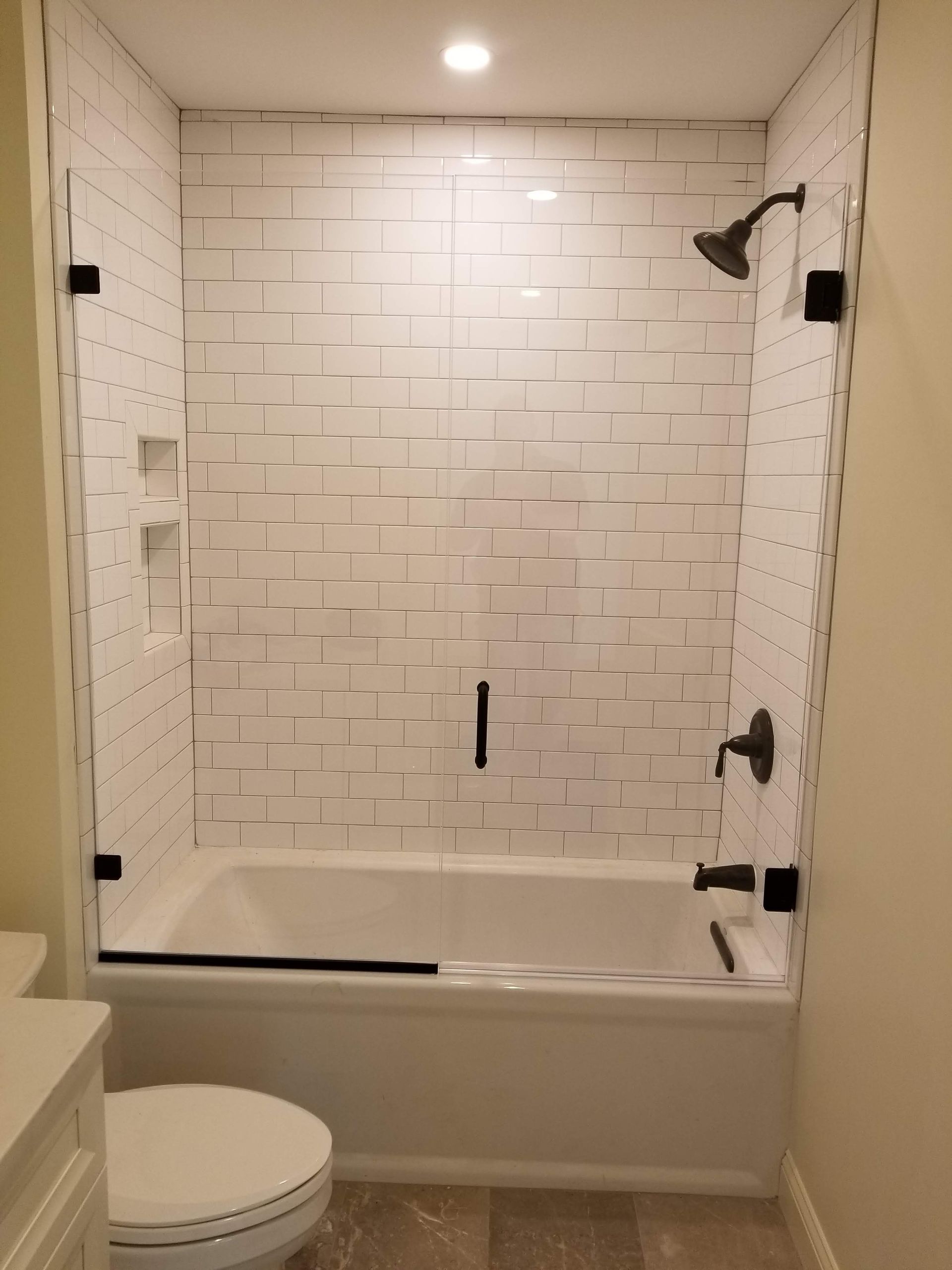 White tiled bathtub and shower enclosure with glass doors, black fixtures, and a toilet.