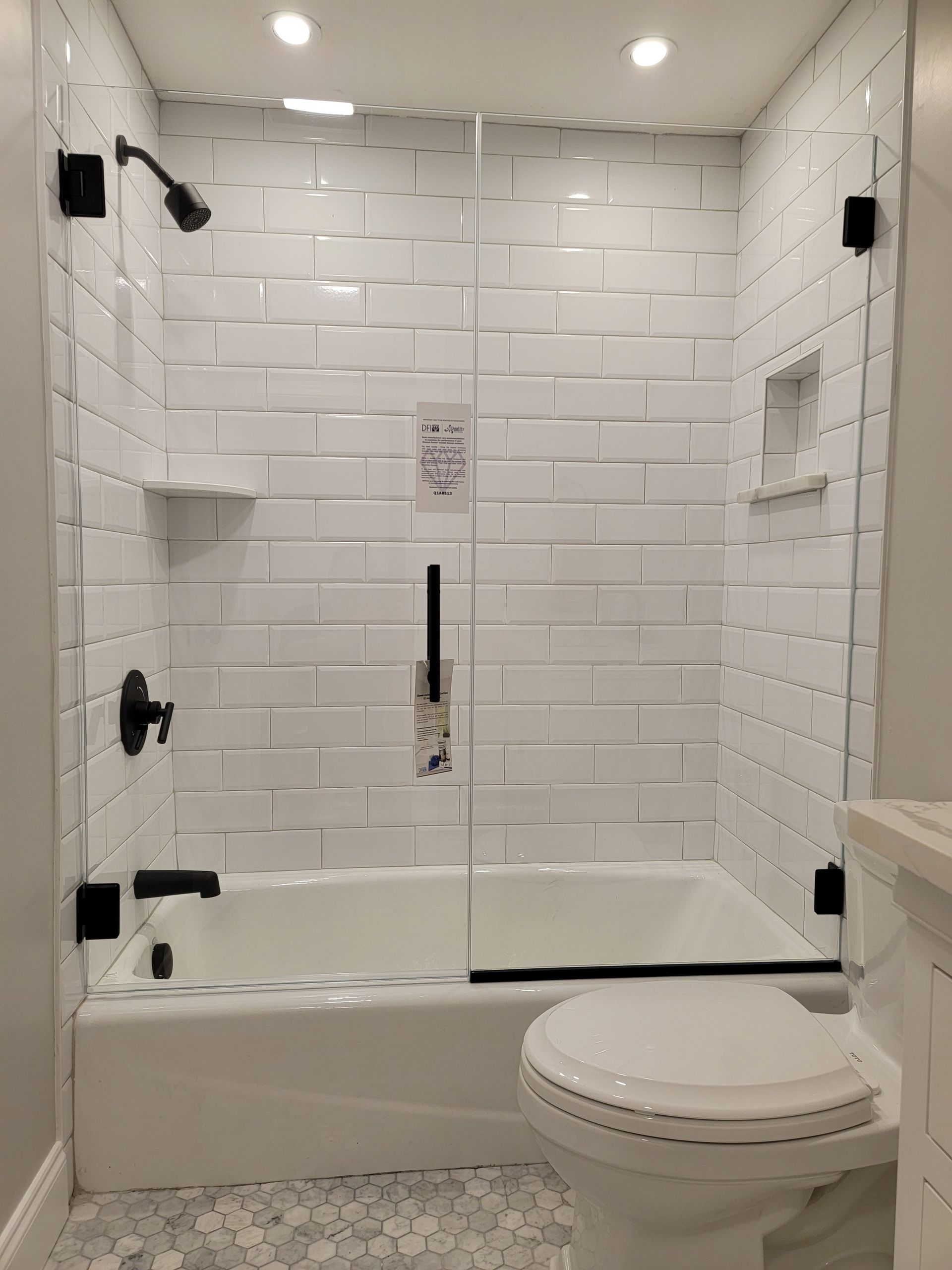 White tiled bathroom with glass shower door, black fixtures, and toilet.