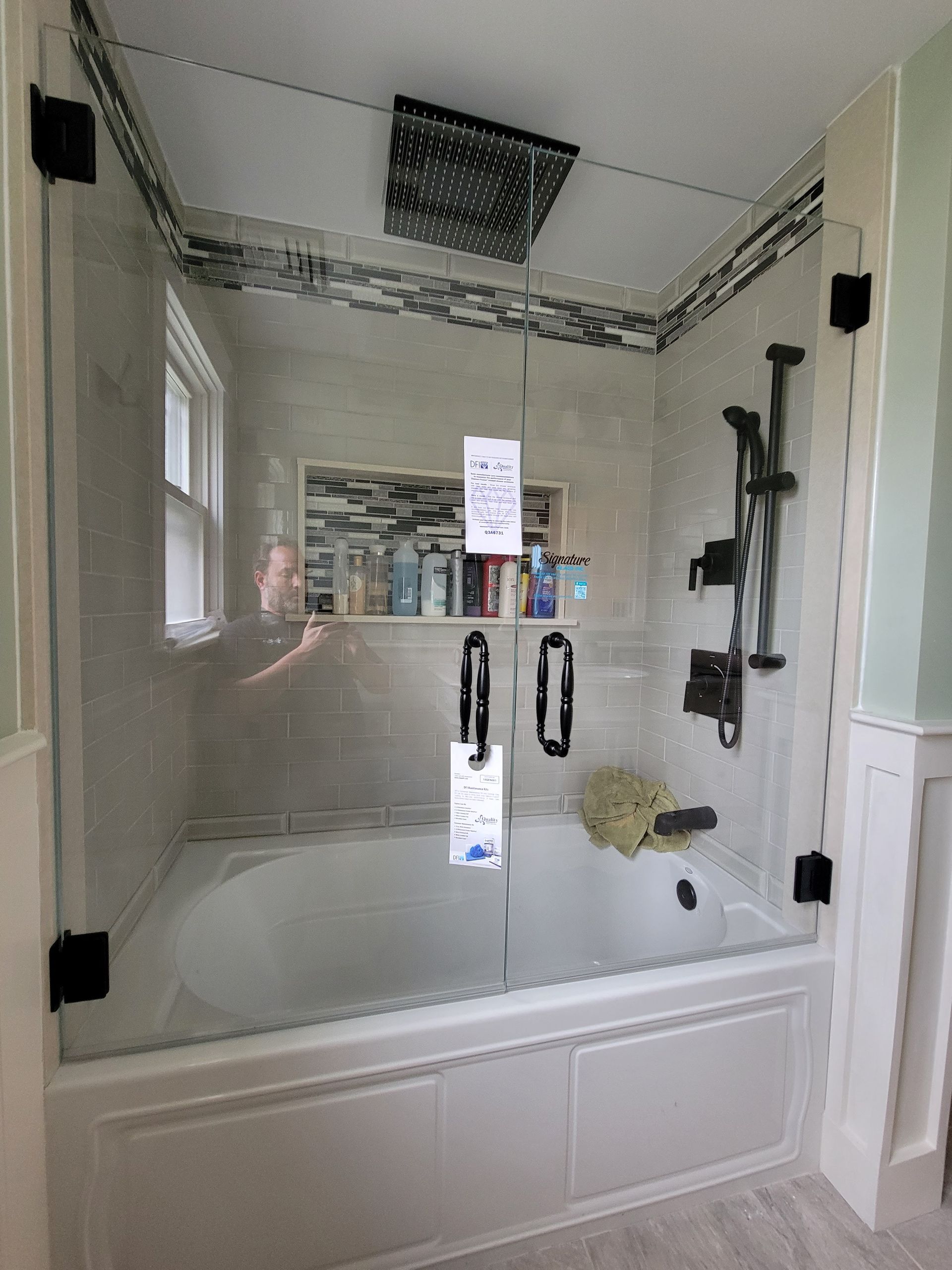 Glass shower enclosure over a white bathtub. Includes a shower head, toiletries, and black hardware.