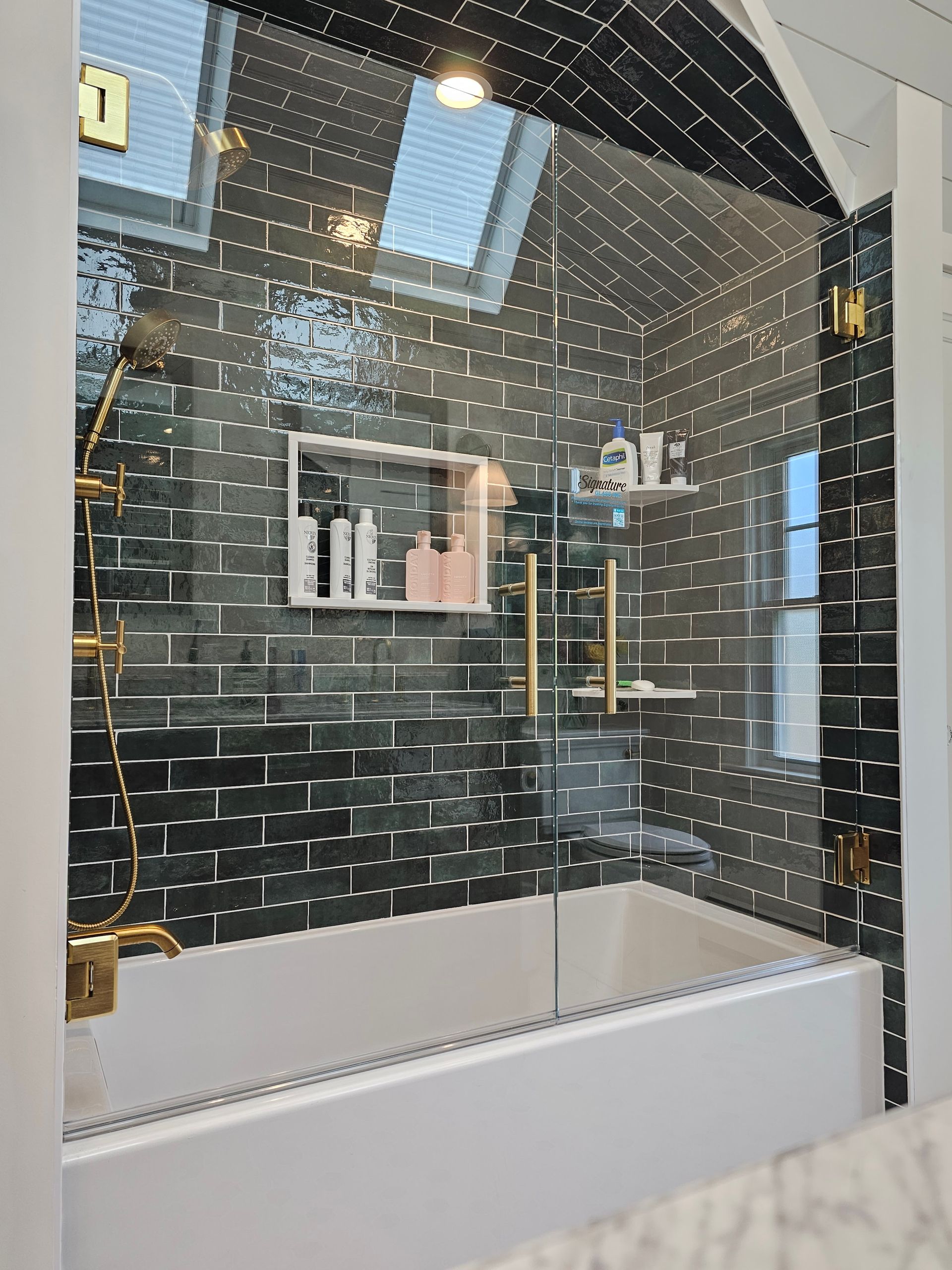 Bathroom with black tiled wall, white tub, gold fixtures, glass shower door.