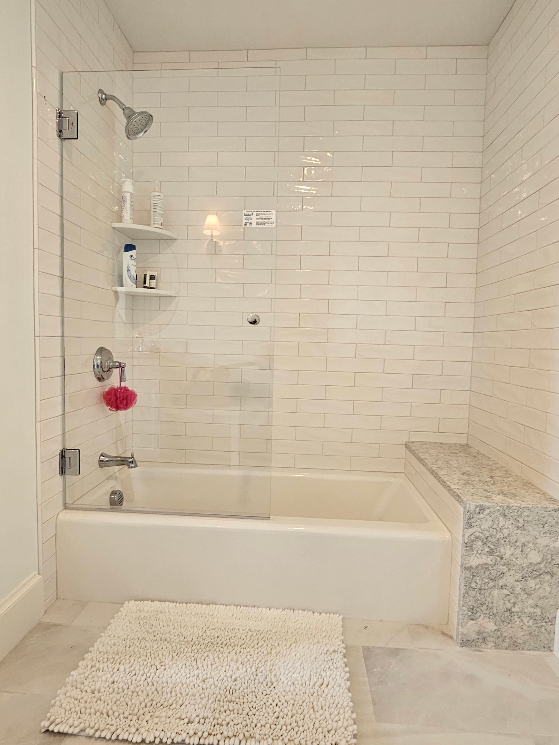White tiled bathroom with glass shower door, tub, bench, and rug.