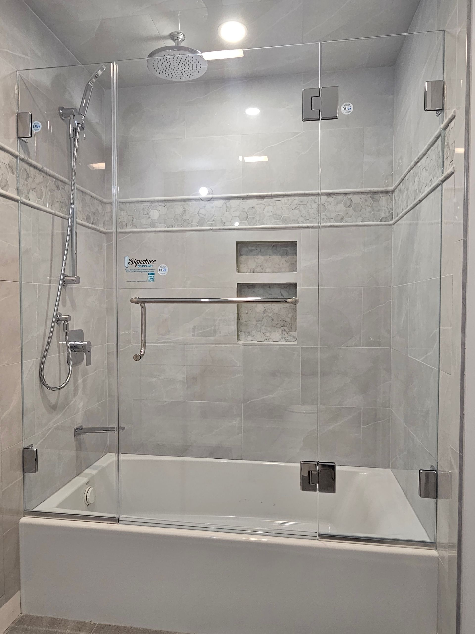 Glass-enclosed shower with a white bathtub and grey tiled walls. Silver fixtures are visible.