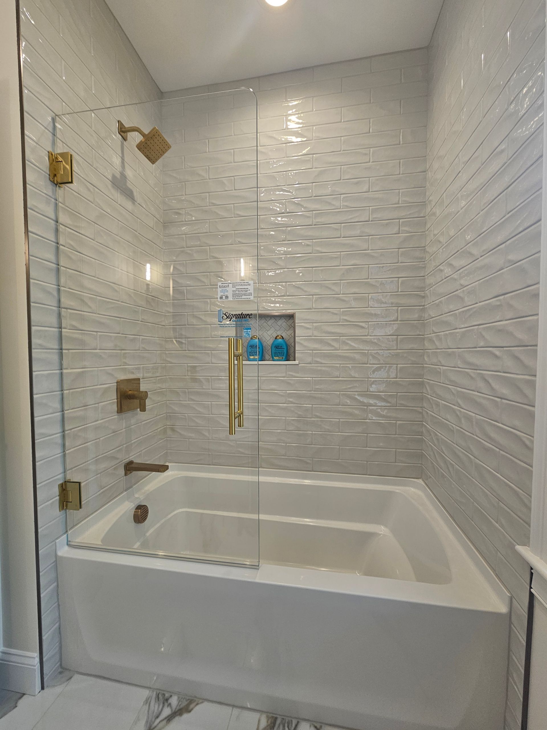 Bathroom with white bathtub, textured white tile, gold fixtures, and glass shower door.