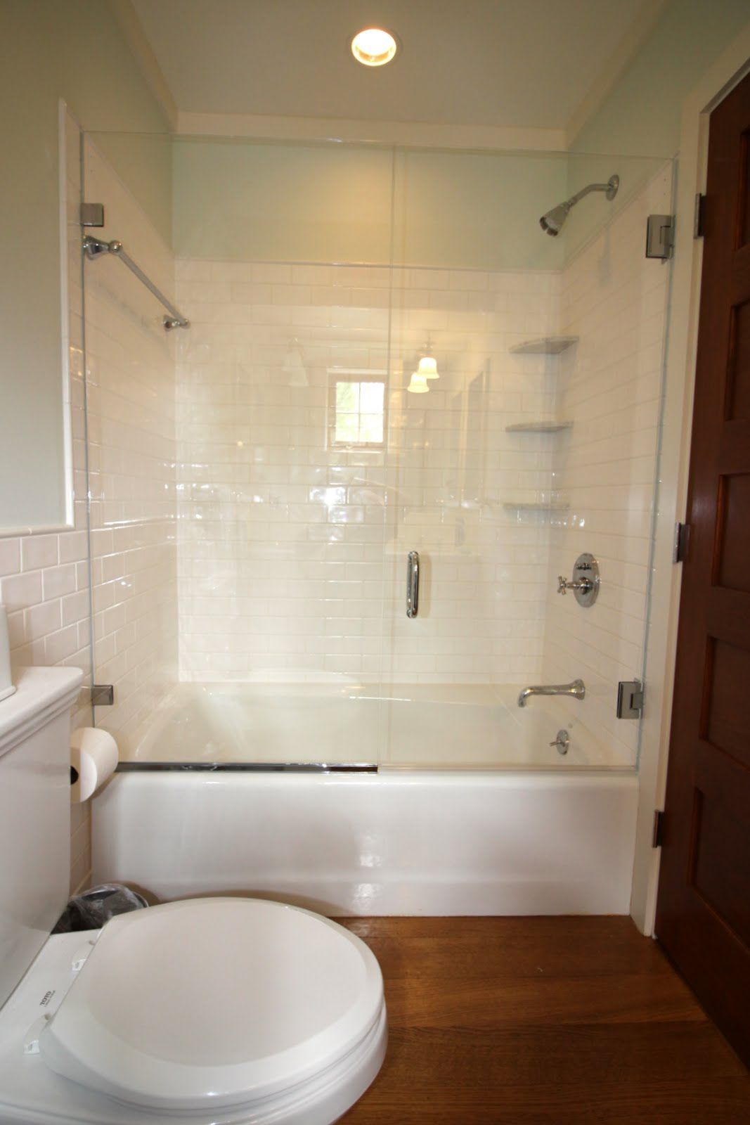 Bathroom with white tub and toilet, clear glass shower doors, wood floor.
