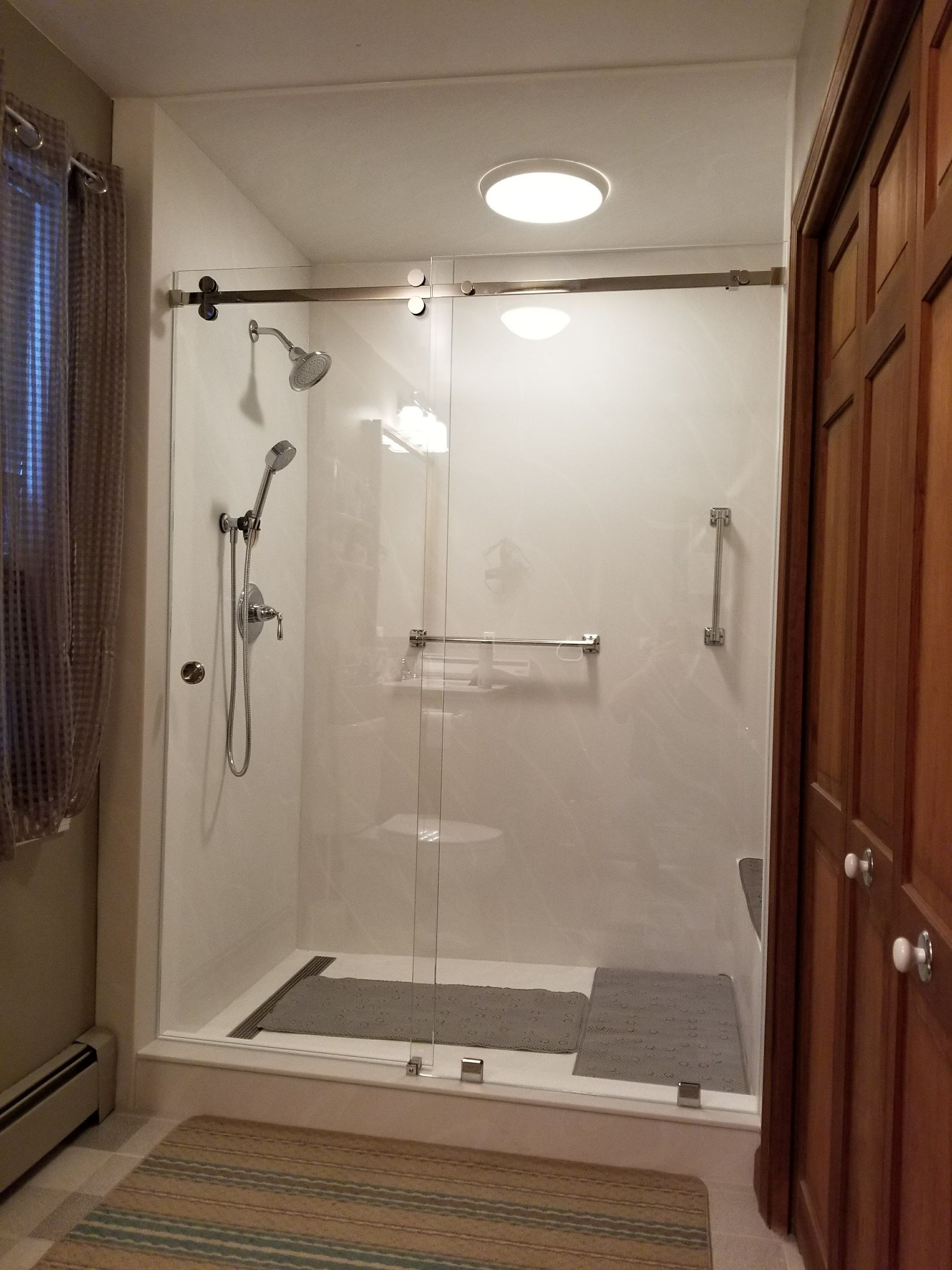 Accessible shower with glass doors, grab bars, and showerheads; light colored walls and floor; rug in front.