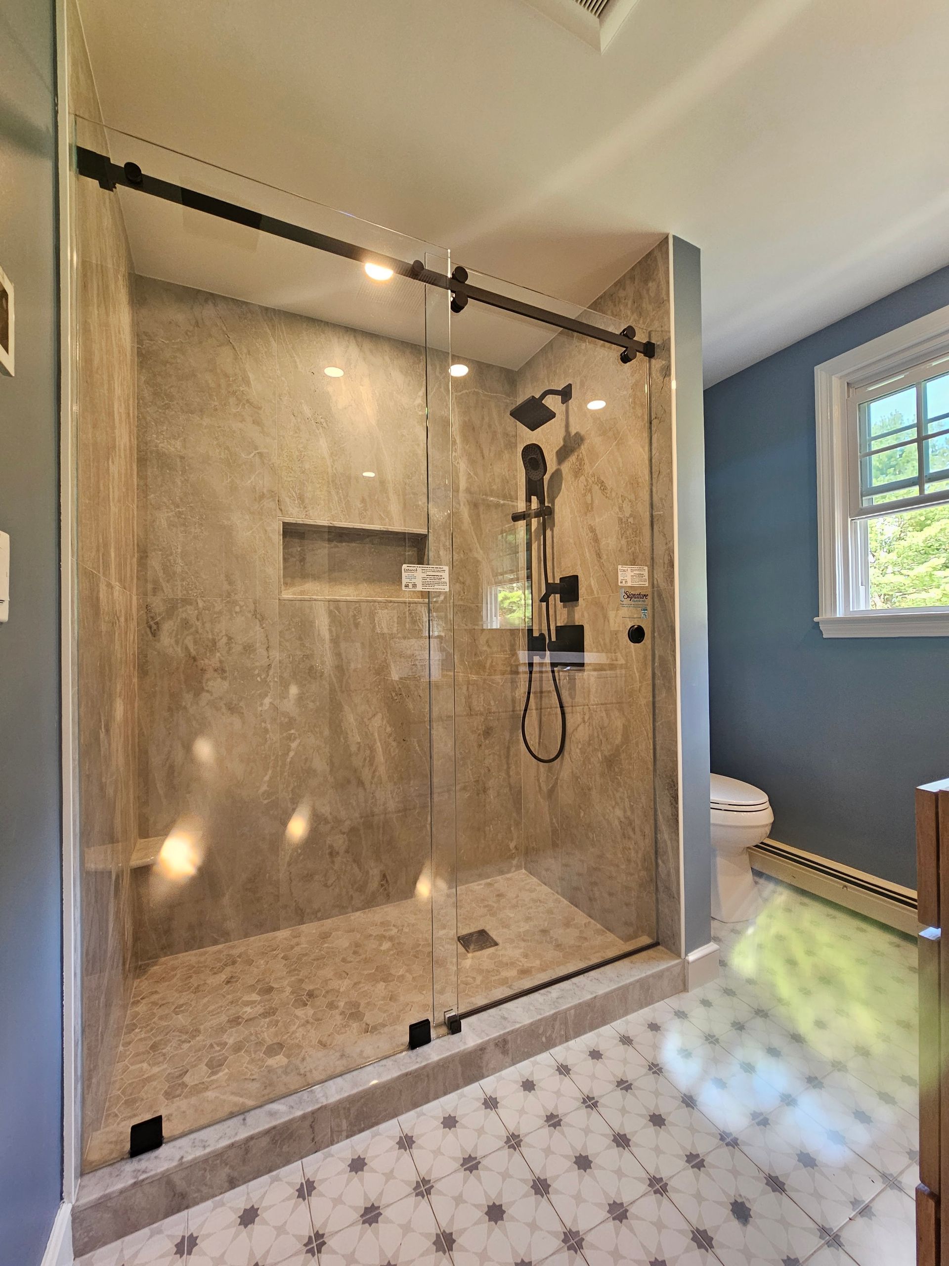 A modern shower enclosure with a sliding glass door and stone tile walls. A black shower fixture is visible.
