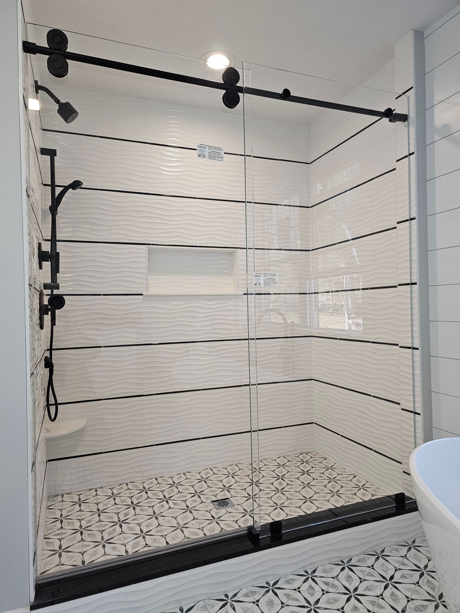 Shower stall with white tile walls, black accents, and glass sliding door. Black and white patterned floor.