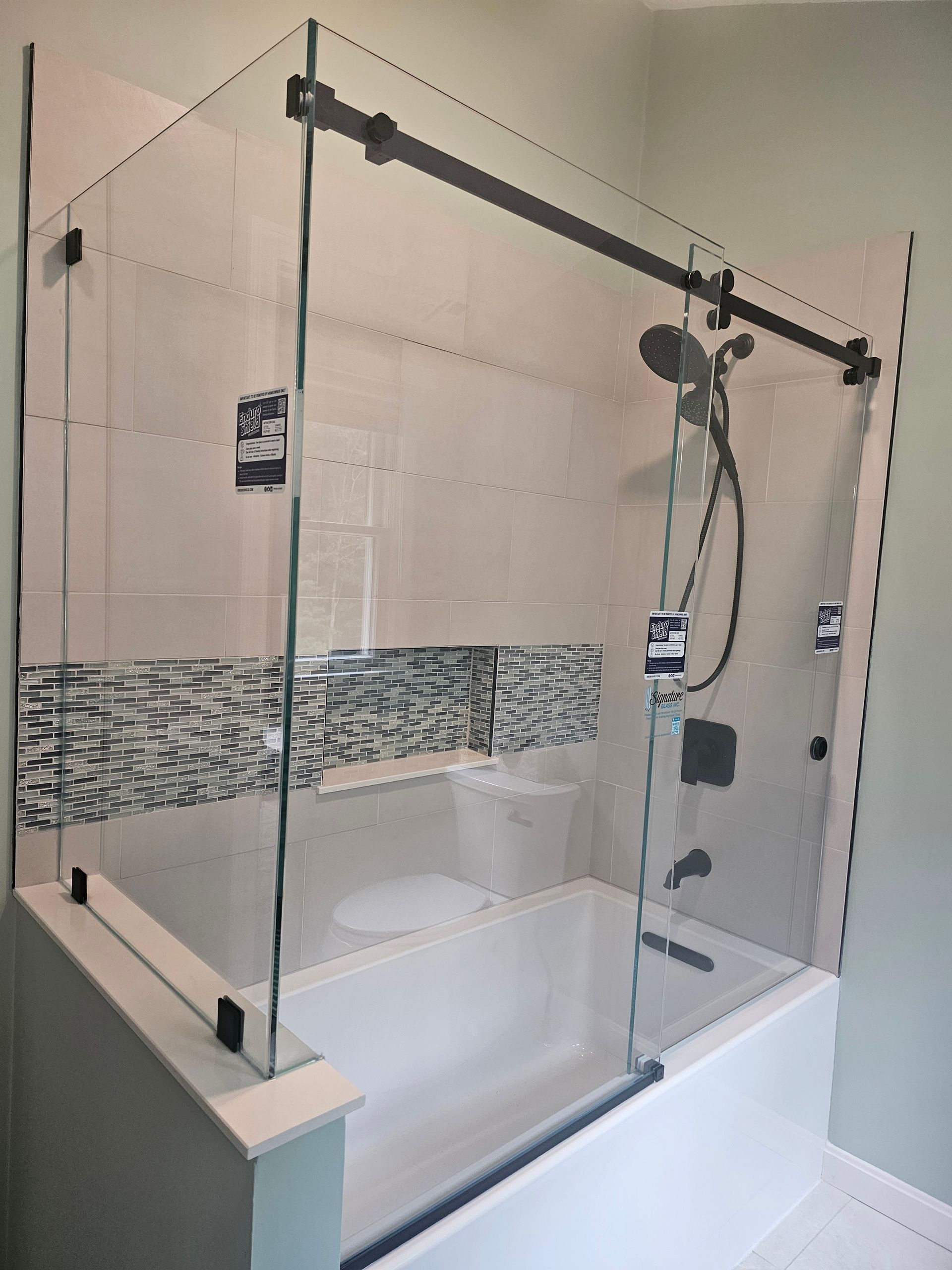 Shower with clear glass doors and black hardware over a white tub with stone tile accent.