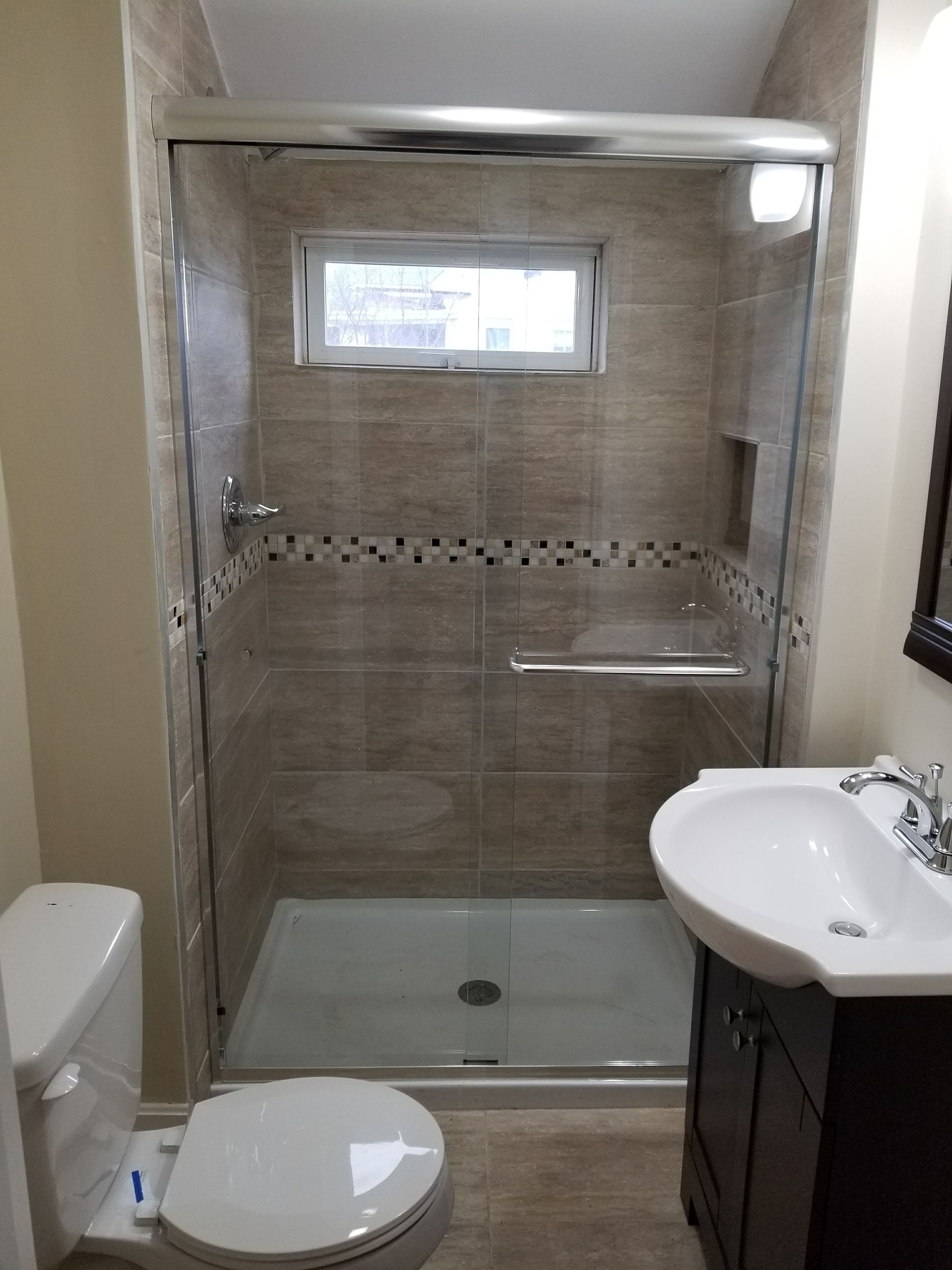 Bathroom with glass shower, toilet, and vanity. Beige tile walls, dark brown cabinet, and a small window.