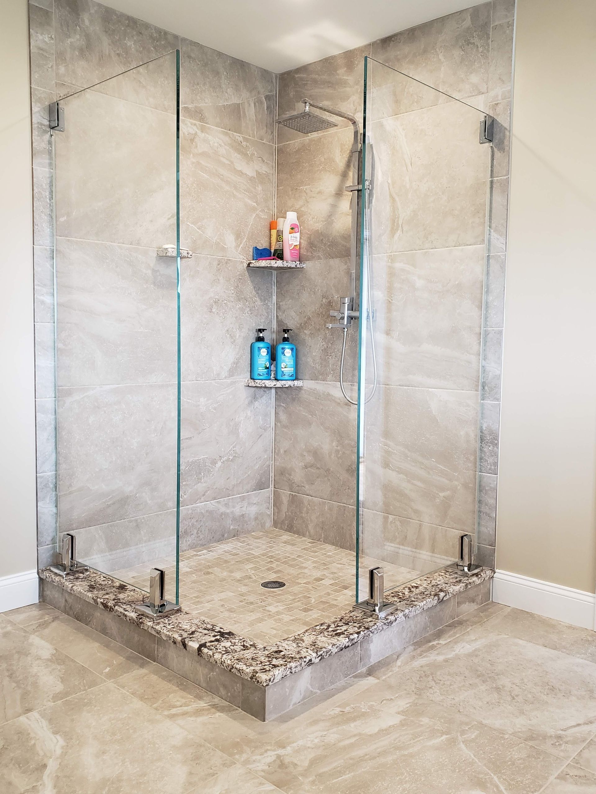Corner shower with glass doors, gray tile walls, pebble floor, and toiletries on a shelf.