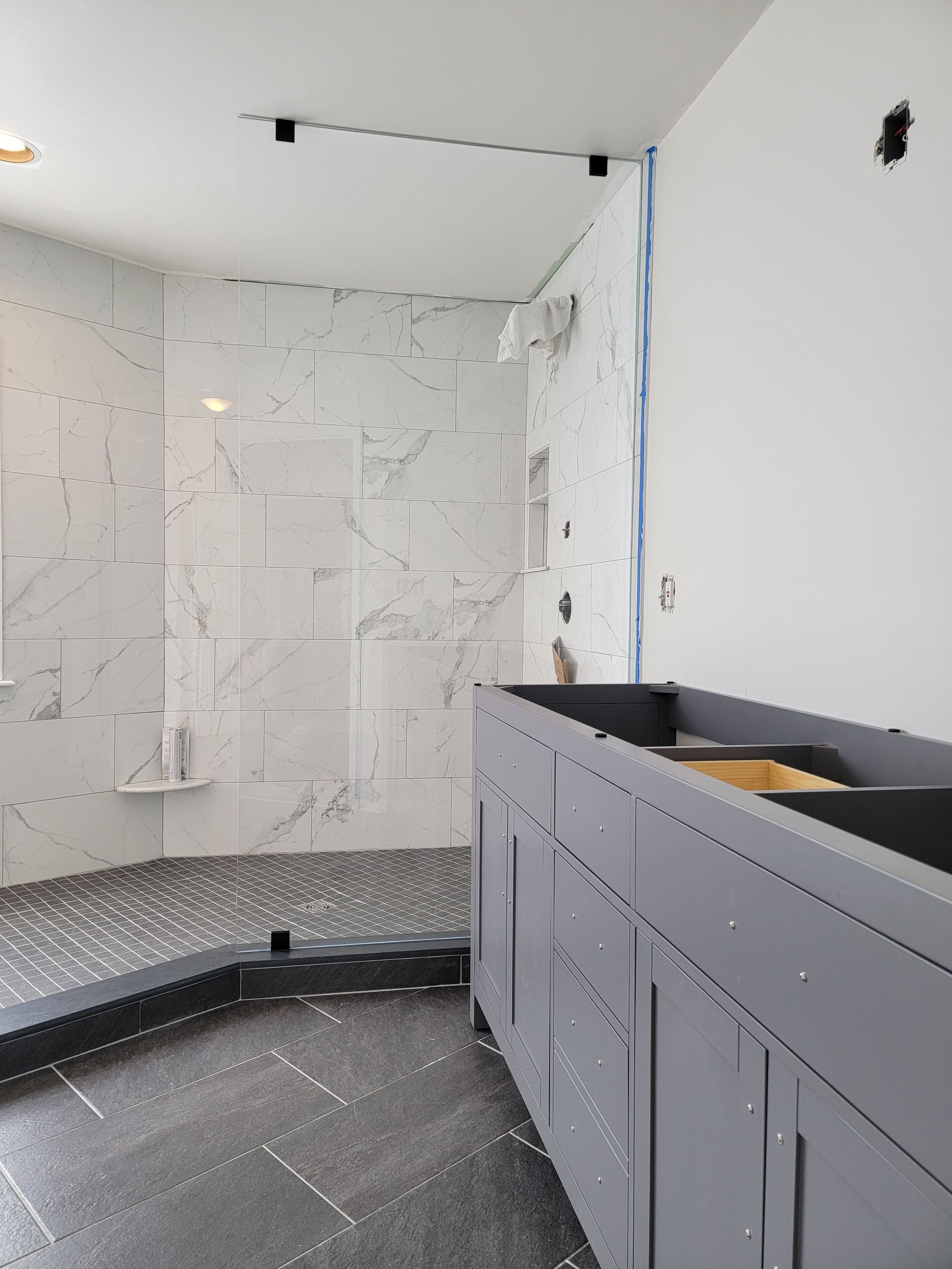 Bathroom under construction with marble-look shower tile, dark floor tile, and gray vanity cabinet.