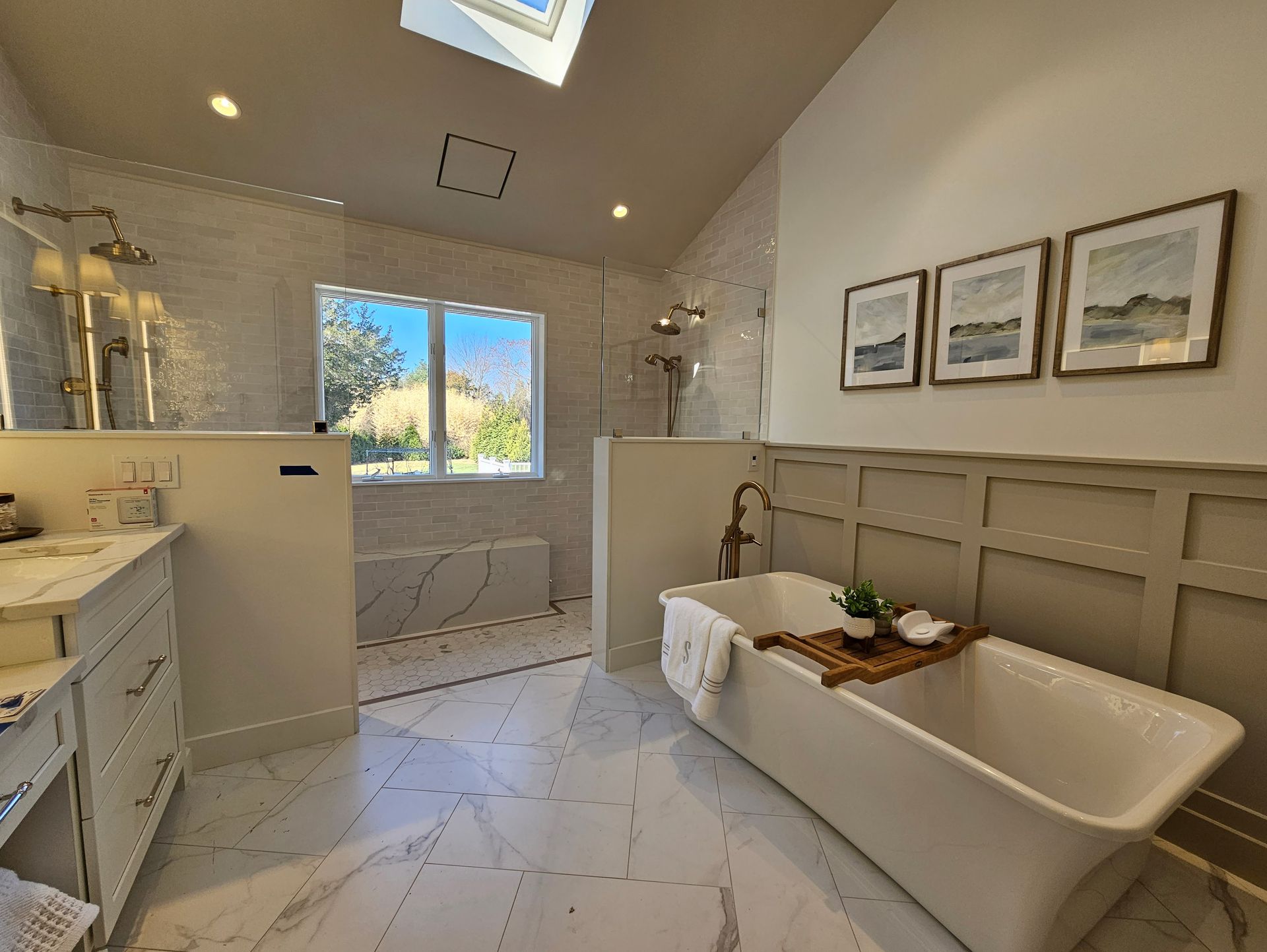 Elegant bathroom with a free-standing tub, white marble tile, shower, and artwork. Natural light from a window and skylight.
