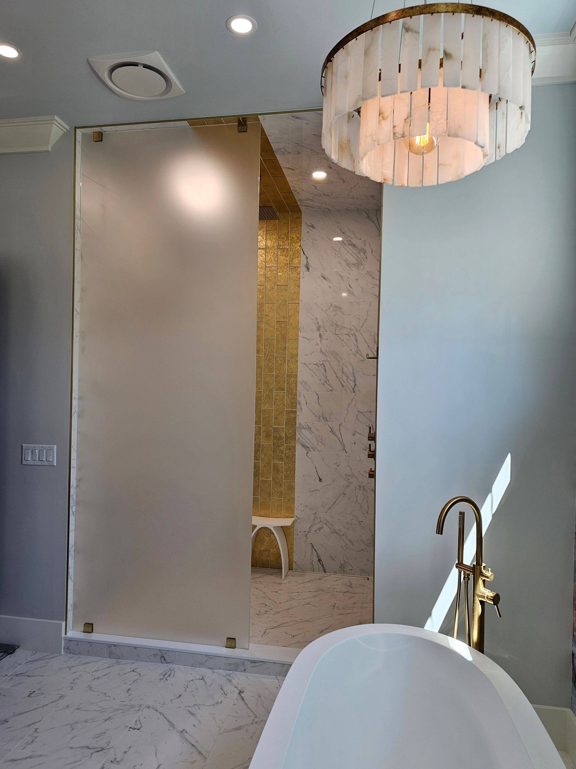 Luxurious bathroom with frosted glass shower door, gold accents, marble, and a chandelier.
