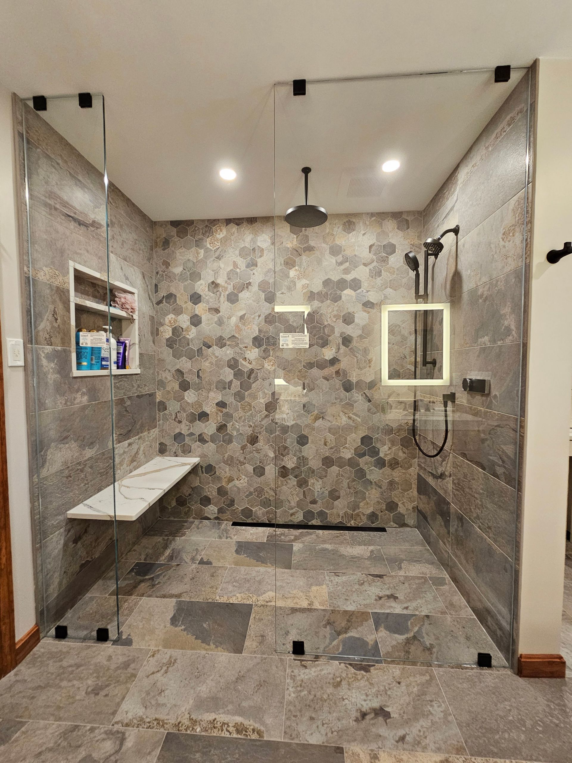Modern shower with gray stone tiles, glass walls, bench, and rainfall showerhead.