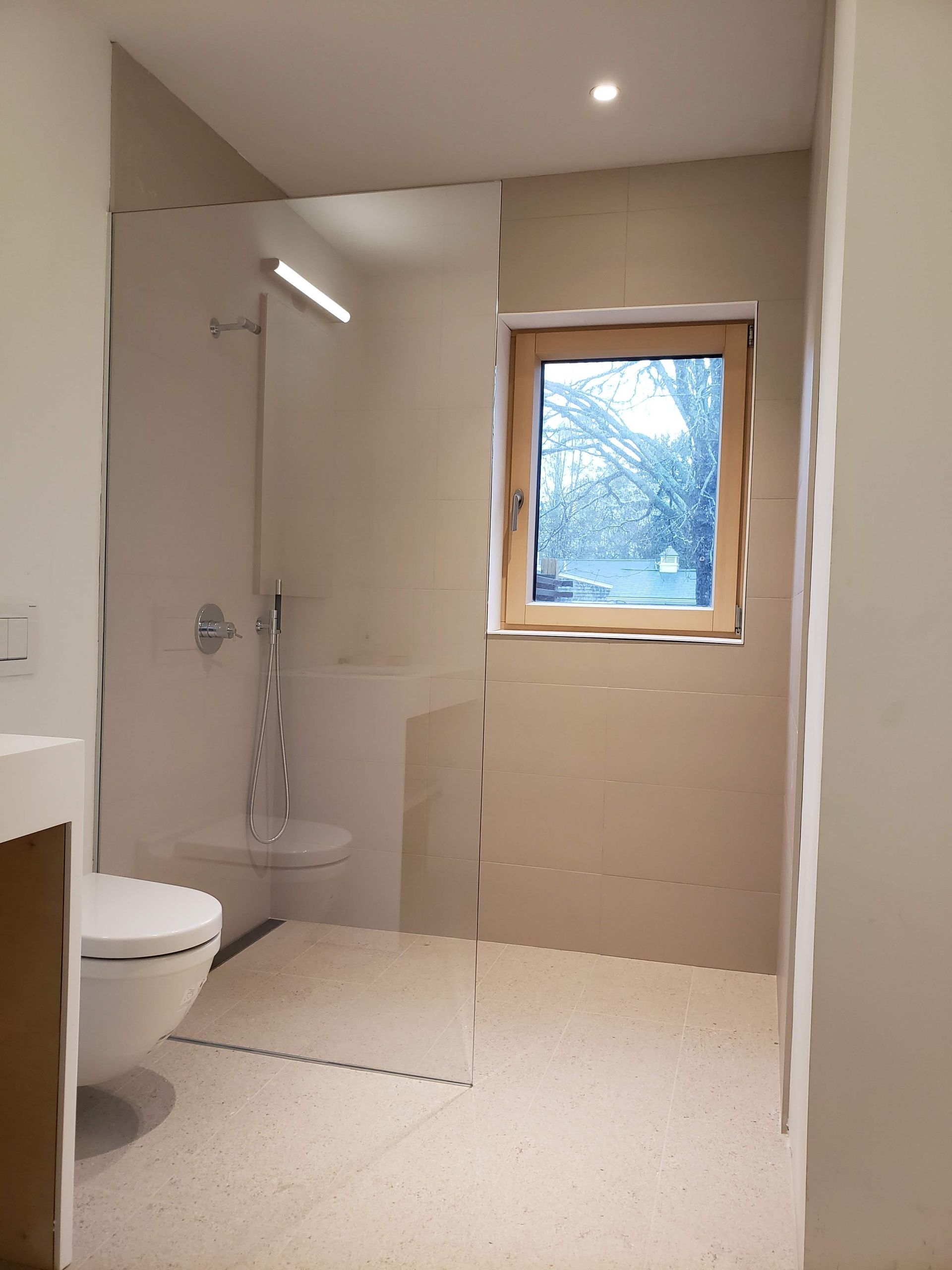 Modern bathroom with glass shower enclosure, toilet, window, and beige tile.