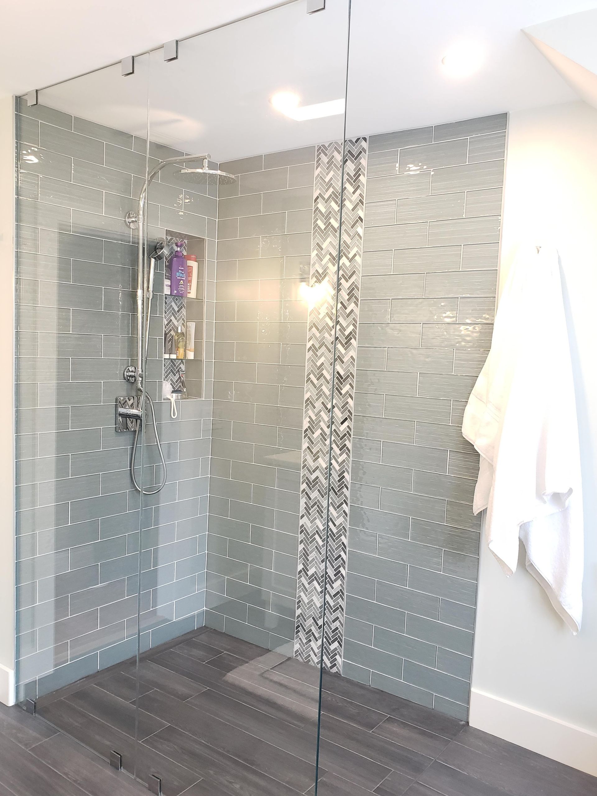 Modern glass shower with gray tiled walls and a decorative strip. Gray floor and white robe on the right.