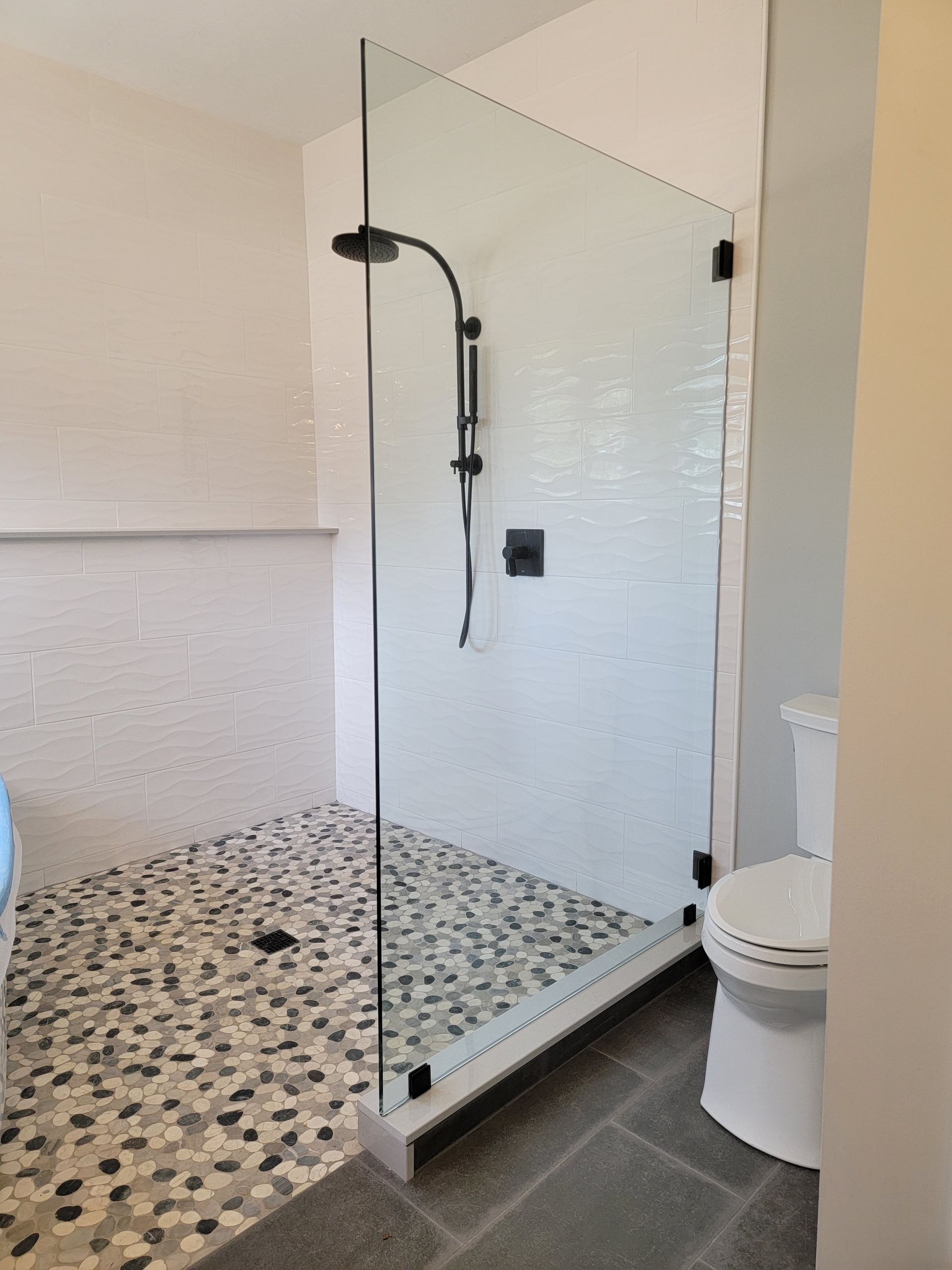 Modern shower with pebble floor, white walls, glass enclosure, and black showerhead. Toilet on the right.