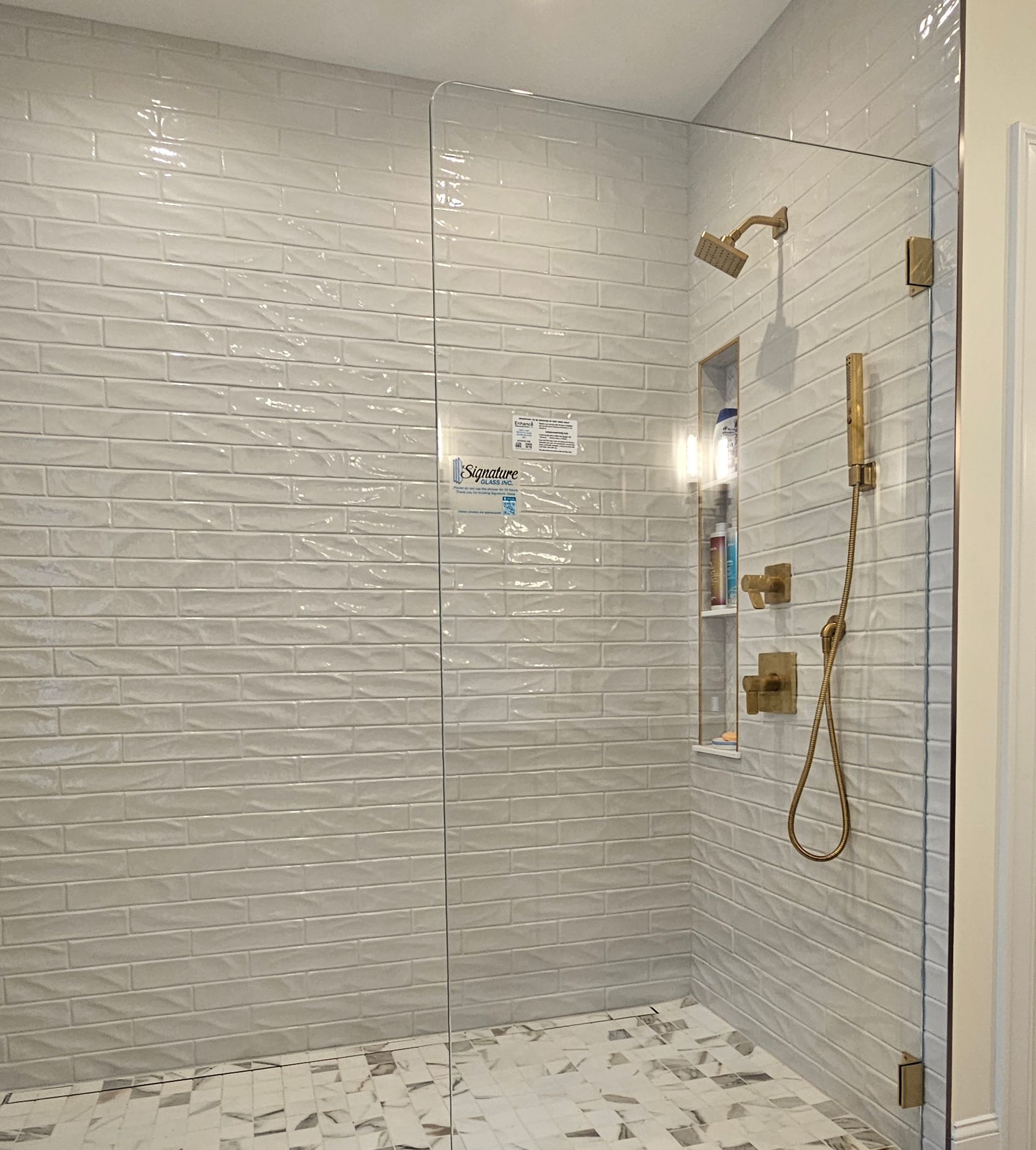 A modern shower with white textured tile walls, glass doors, and gold fixtures.