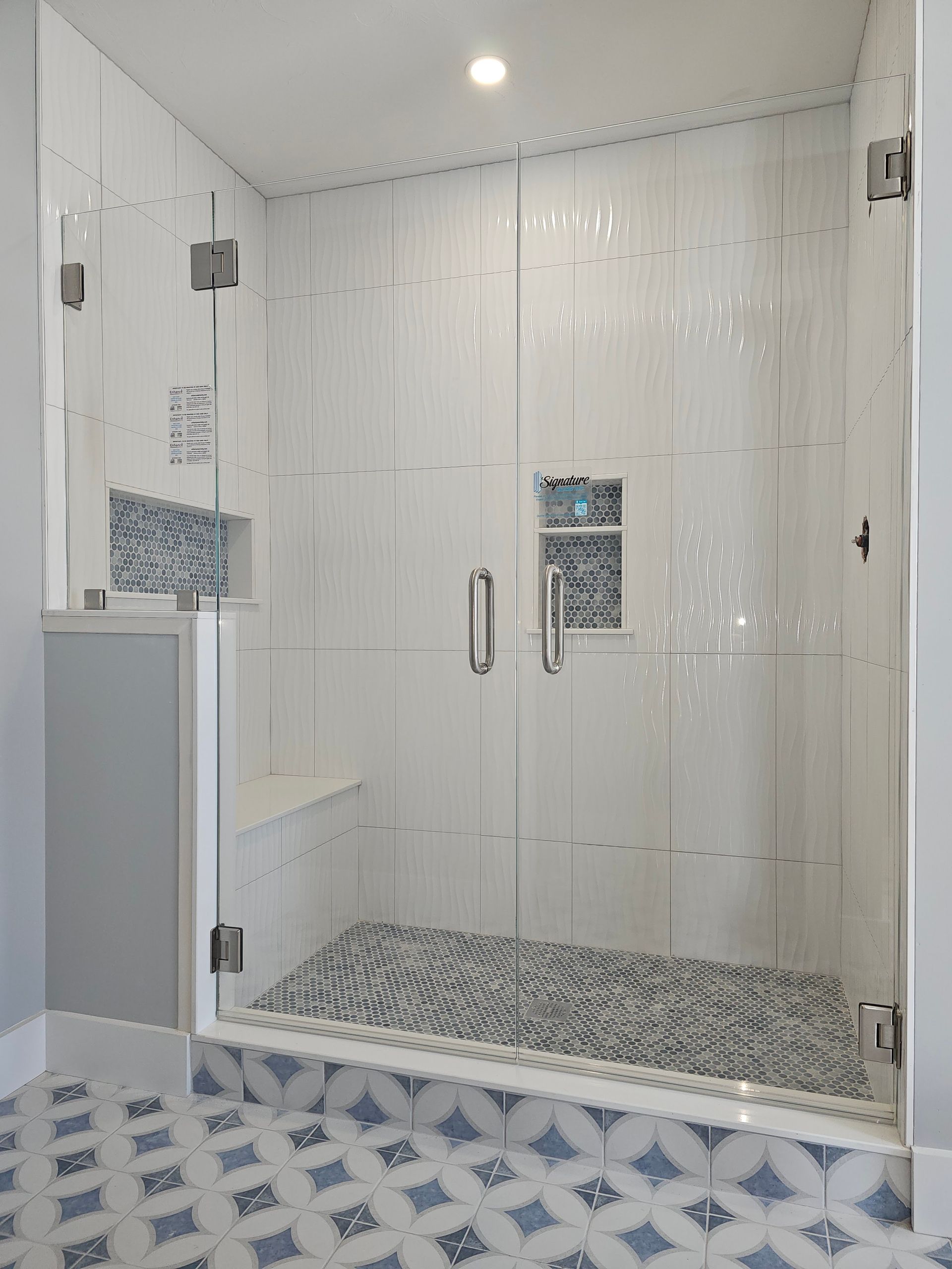 Glass-enclosed shower with white textured tiles, mosaic floor and accent, and blue and white patterned floor tiles.