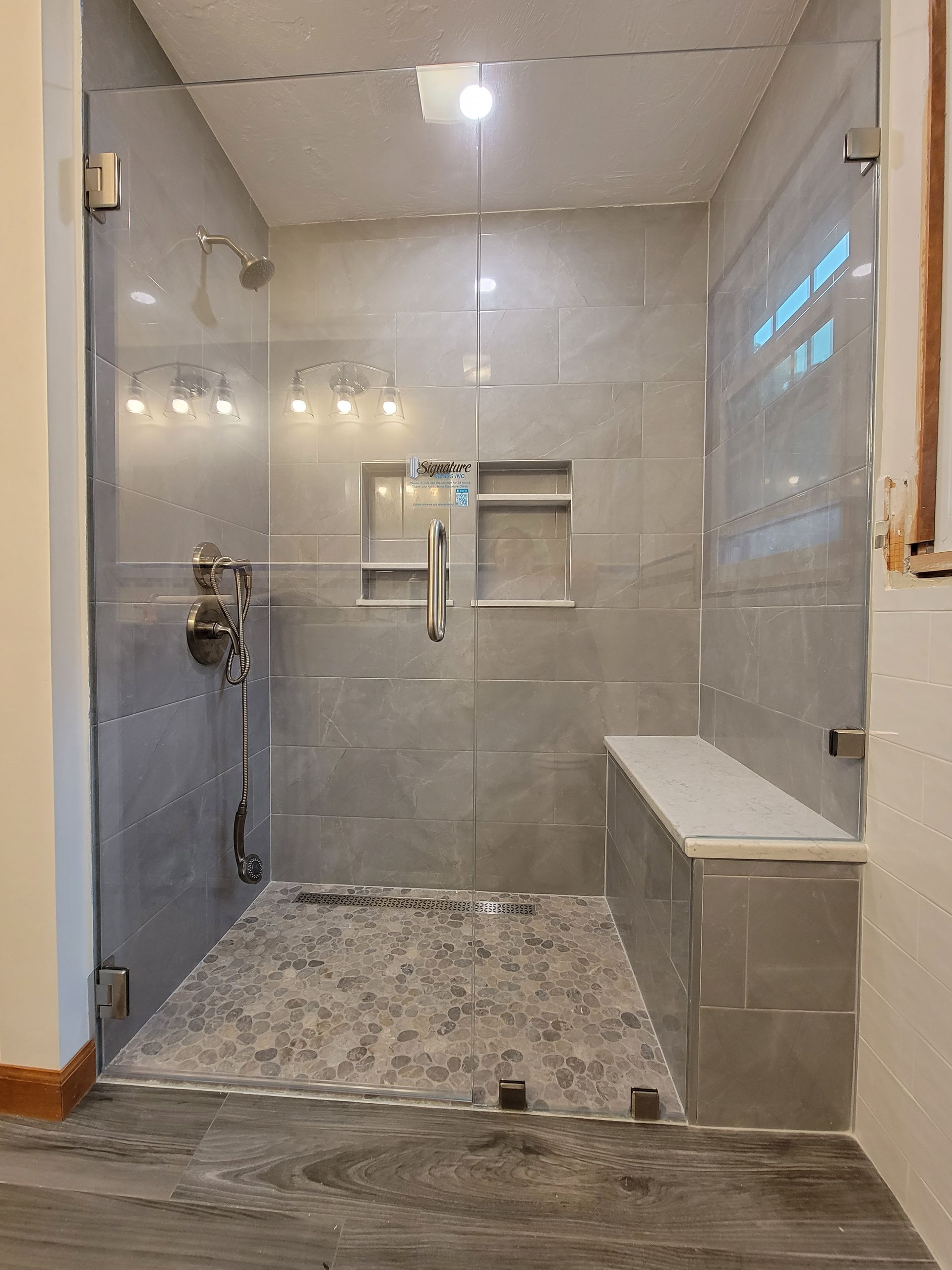 Glass-enclosed shower with gray tiled walls and floor, a bench, and recessed shelves.